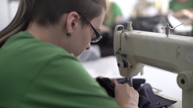 Young Caucasian girl sitting at sewing machine and stitching factory textile, profile of face close up, shallow depth of field, young female person tailor portrait, interior scene