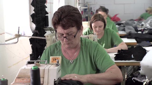 Portrait of mature Caucasian female tailor with glasses during sits for the sewing machine and tailoring, group of women working in the blurred background, shallow depth of field, interior scene