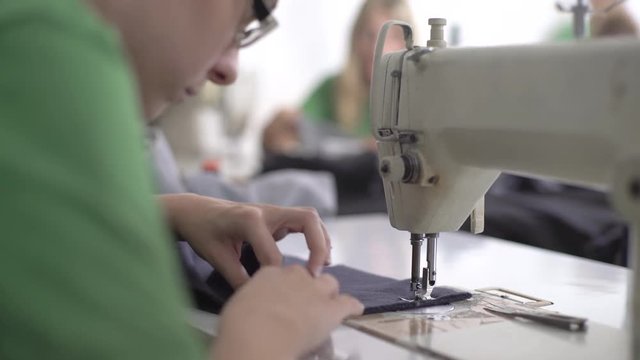 Young Caucasian female person in t-shirt blurred sitting using sewing machine to stitching factory textiles, shallow depth of field, close up, interior scene