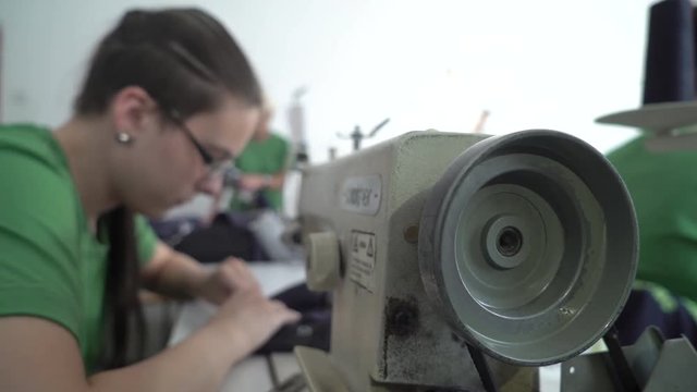 Sewing machine in work process close up in focus, young female tailor stitching fabric textiles in the blurred background, shallow depth of field, interior scene