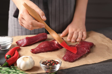Woman applying oil onto raw steak with silicone brush in kitchen