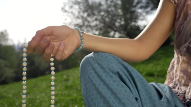 Woman, lit hand close up, counts Malas, strands of wooden beads used for keeping count during mantra meditations. Buddhism. Girl sitting in the park at summer. 4k