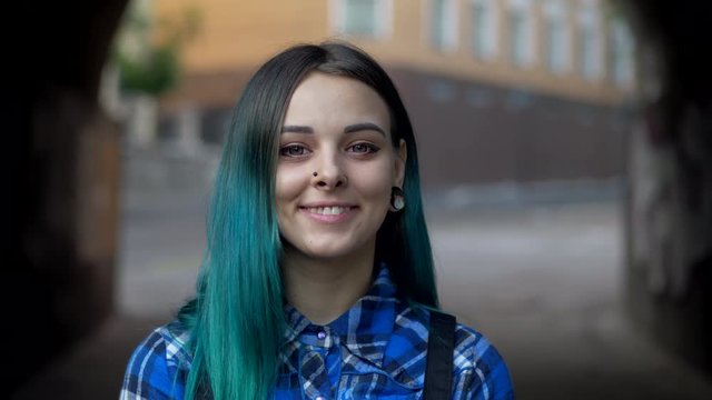 Street trendy or hipster girl with blue dyed hair. Woman with piercing in nose, violet lenses, ears tunnels and unusual hairstyle stands in city. 4k