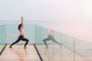 Asian women play yoga on a wooden deck by the sea in the morning. With reflection in the mirror.