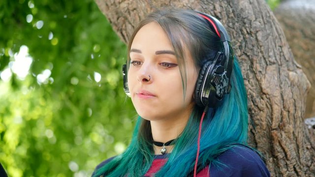 Street punk or hipster girl listening music with black headphones. Teen girl with blue colorfulunusual dyed hair,piercing in nose, sitting on big tree in park . 4k