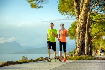 Poster Junges sexy Paar läuft die Promenade entlang. Sie machen ihr Sport-Workout im wunderschönen Sonnenuntergang am Strand. buntes Kleid, Bäume, Wasser, Berge und ein unglaublich blauer Himmel. © Merpics