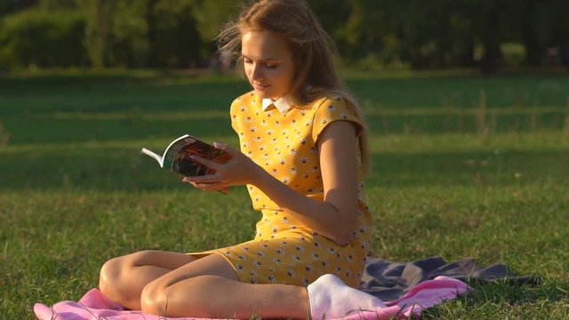 Slow motion. A young blonde girl with European outfits sitting in a warm sunny day on a meadow in the park. read book. a woman relaxes in the rays of the setting sun and remuved her hair off her face