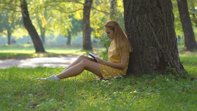 Slow motion. A young blond girl in a warm sunny day at a picnic. She is dressed in a bright yellow dress. green grass . holds in his hand a book and reads. rest in the park. the woman is sitting near