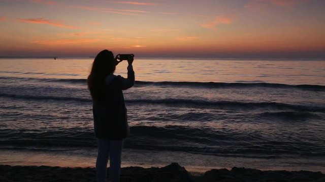 Young woman taking photos with her smartphone, on the beach, at sunrise