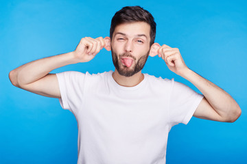 Funny young Caucasian man grimacing making mouths, sticking out his tongue at camera, making ears...