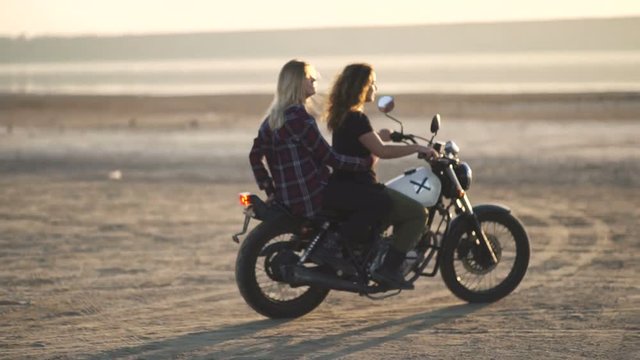 beautiful young woman motorcyclist with his girlfriend riding a motorcycle in a desert on sunset or sunrise. Girlfriends on a bike. Slow motion
