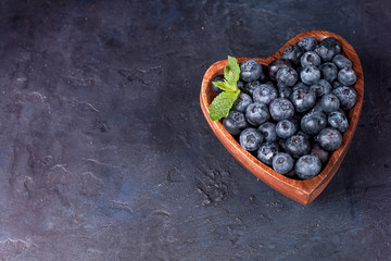 Fresh blueberries with a leaf of mint in the heart-shaped bowl on a dark blue background