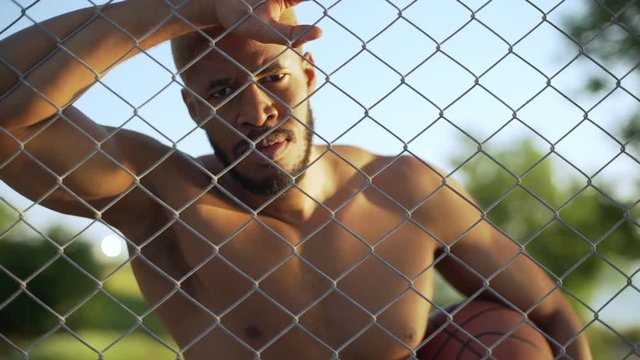 naked young guy with blond hair with basketball under his arm leaning on chain link fence and looking at camera in slow motion