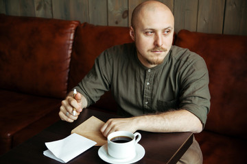 Young serious fashionable man sitting alone and putting down his thoughts in his notebook in loft-styled cafe. Former factory building, mixed lighting.