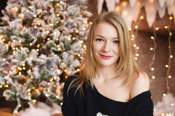 Happy young lady with blonde hair sitting by the fireplace near the Christmas tree. New year concept.