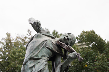 Bronze statue of musician. Violinist and fiddler plays the violin. Trees and white sky. Sculpture as aged and green. Figure has covered eyes by scarf - sensuality
