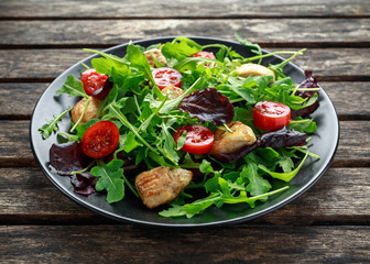 Fresh salad with chicken breast, arugula, nuts and tomatoes on black plate in a wooden table.