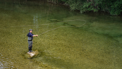 Angler mit Wathose und Fliegenrute im Wasser beim Angeln bei Sonne im klaren Fluss stehend und werfend