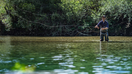 Fototapeta na wymiar Angler mit Wathose und Fliegenrute im Wasser beim Angeln bei Sonne im klaren Fluss stehend und werfend