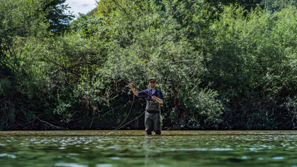 Fototapeta na wymiar Angler mit Wathose und Fliegenrute im Wasser beim Angeln bei Sonne im klaren Fluss stehend und werfend