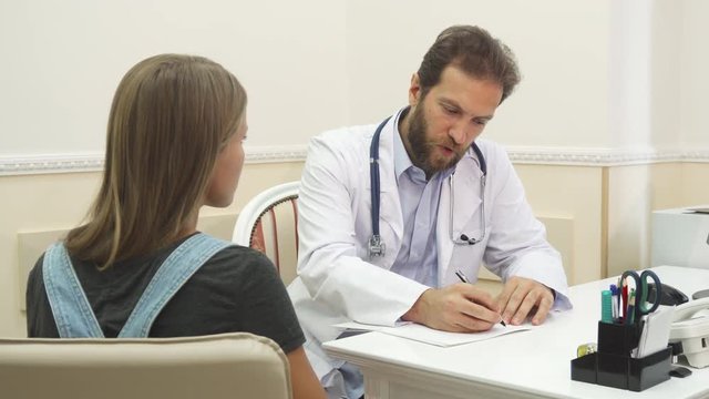 The doctor is writing recommendations to his patient. The accent is made on the gestures of the doctors hands. The girl comes to the therapist. They are sitting in doctors office