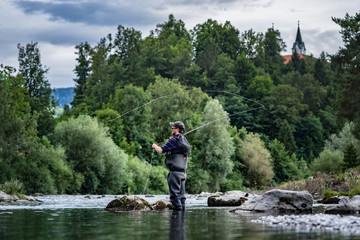 Angler mit Wathose und Fliegenrute im Wasser beim Angeln bei Sonne im klaren Fluss stehend und...