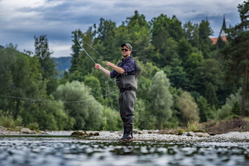 Angler mit Wathose und Fliegenrute im Wasser beim Angeln bei Sonne im klaren Fluss stehend und...