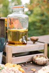 food, refreshment, drinks concept. in the big transparent glass jar with special silver spigot there is yellow lemonade nearby with different tasty snackes like nuts