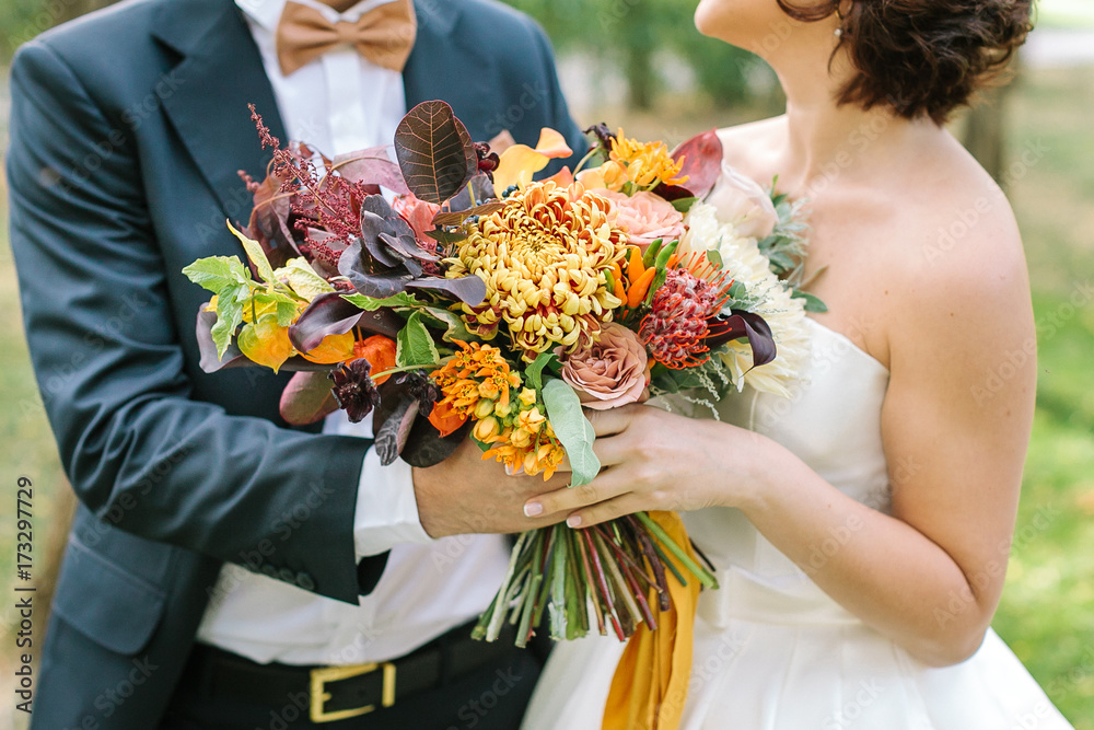 Wall mural autumn, occasion, love concept. fiance dressed in magnificent black suit and creamy bow tie and his bride with delicate shoulders holding togethere wedding bouquet of fall shades