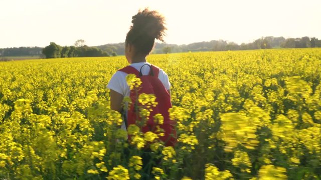 4K video clip of beautiful happy mixed race African American girl teenager female young woman hiking with red backpack in field of rape seed yellow flowers