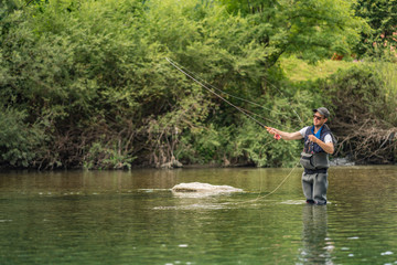 Angler mit Wathose und Fliegenrute im Wasser beim Angeln bei Sonne im klaren Fluss stehend und werfend