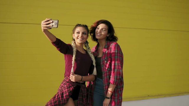 Two best friends girls having fun and making selfie standing by the yellow wall. Two hipster girls taking selfie photos with smart phone and smiling. Slowmotion shot