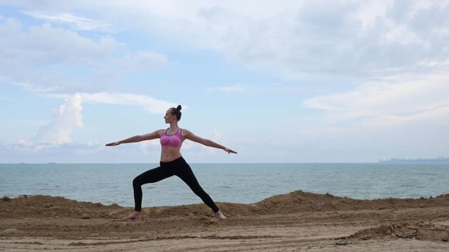 Girl practices yoga fitness exercise near the ocean beach in warrior pose 4k