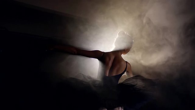 ballerina with hair in a bun is dancing on a scene, against bright light of soffit and smokescreen