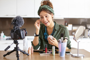 Attractive young woman beauty fashion blogger using sponge while applying powder, talking about cosmetics and filming make up tutorial to attract more followers to her blog on social networks