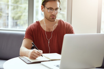 Hardworking young bearded man wearing hat and glasses studying via Internet, listening to business webinar in earphones, looking at laptop screen and writing down important notes in leather notebook