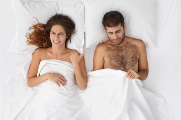 Indoor shot of unhappy frustrated bearded Caucasian man looking under white blanket at his penis, worried because of erectile dysfunction or small size, his wife pointing at him, having disgusted look