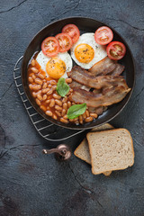 Metal frying pan with english breakfast including bacon, fried eggs and beans in tomato sauce. Above view on a dark grey textured background, vertical shot