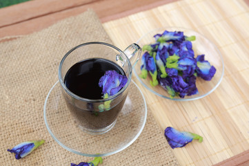 Butterfly pea juice in transparent glass and flowers on sack-wood mat background.