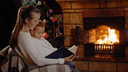 Mother with little daughter reading book near fireplace and christmas tree