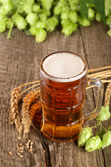 glass of foamy beer with hop cones and wheat on old wooden background