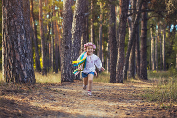 A little girl in a wreath with ribbons is running happily through the summer forest.