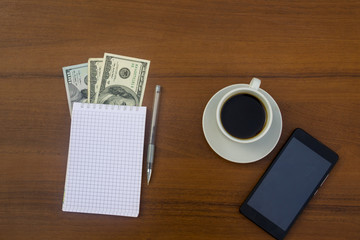 Cup of coffee, smartphone, dollars, notepad and pen on wooden desk