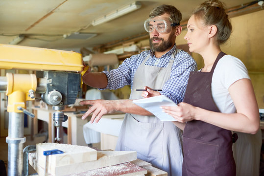 Portrait of bearded carpenter explaining operating process of drilling machine to his female assistant in woodworking shop