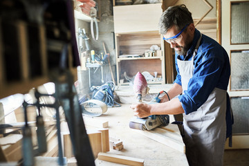 Portrait of mature bearded carpenter polishing piece of wood in workshop against window while making furniture