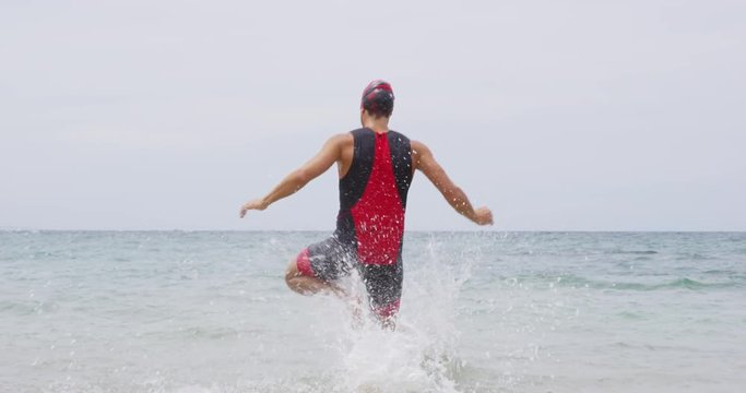 Triathlon swimming - male triathlete swimmer running into ocean for swim. Fit man starting swimming doing freestyle crawl strokes in professional triathlon suit training for ironman.