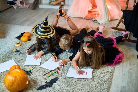 Concentrated little friends in beautiful costumes lying on carpet and drawing on paper imaging Halloween characters at kids party