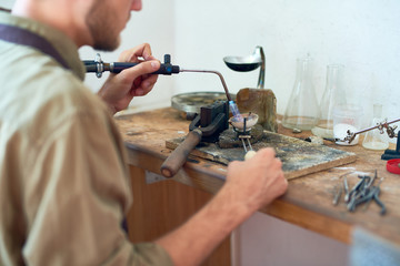 Back view of young man holding small gas torch and melting tools while working with metal at shabby wooden workstation