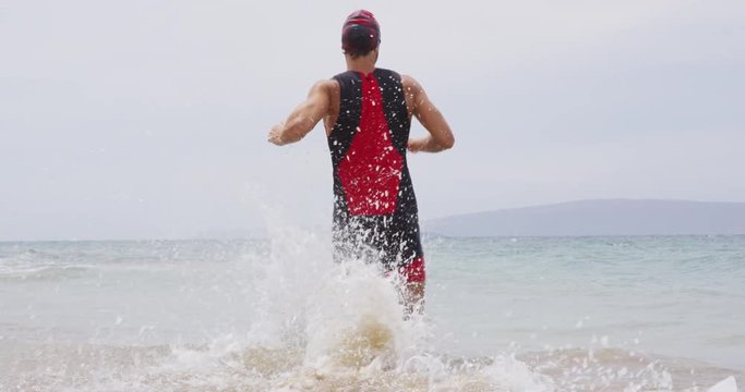Triathlon swimming - male triathlete swimmer running into ocean for swim. Fit man starting swimming freestyle crawl strokes in professional triathlon suit training for ironman on Hawaii.