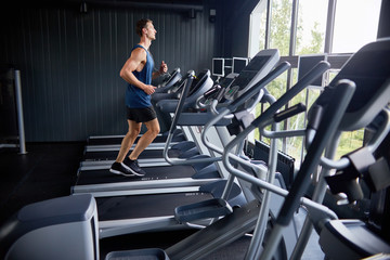 Profile view of handsome young man in sportswear enjoying picturesque view from panoramic window while running on treadmill at modern sports center